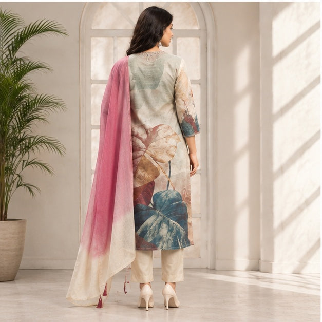 A woman stands with her back to the camera wearing a three-piece printed salwar kameez suit with a pink dupatta, in a bright room with arched windows.