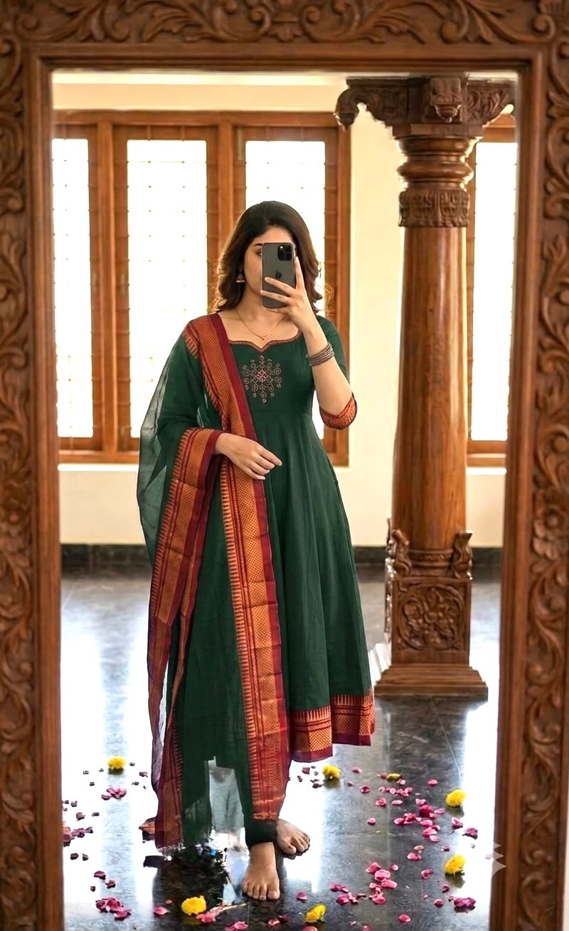 A woman takes a mirror selfie wearing a dark green salwar kameez suit with a red and gold patterned dupatta, standing in front of a large ornate wooden mirror.