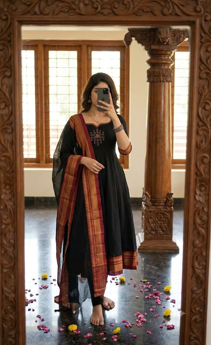 A woman takes a mirror selfie wearing a black kurta with a red and gold patterned dupatta, standing in a room with wooden columns and windows.
