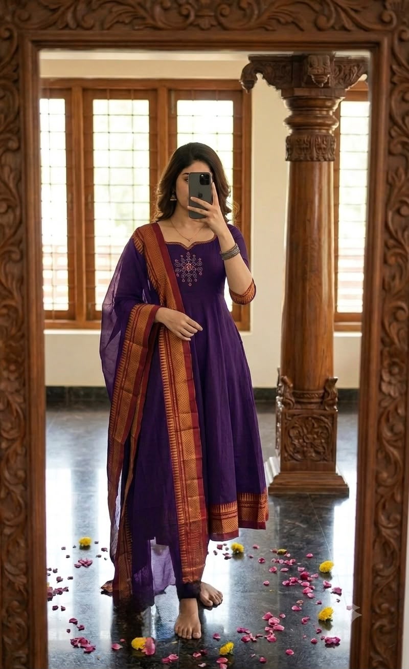 A woman takes a mirror selfie wearing a purple anarkali kurta with a matching dupatta featuring orange and gold borders, set in a room with a carved wooden pillar and window.